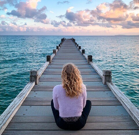 Woman sat on a wooden pier reaching out to the sea, meditating