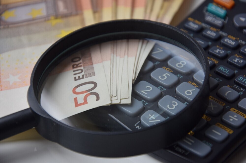 Image of a magnifying glass magnifying 50 Euro Notes place on a scientific calculator to signify the soft cost of buying a home and land. 