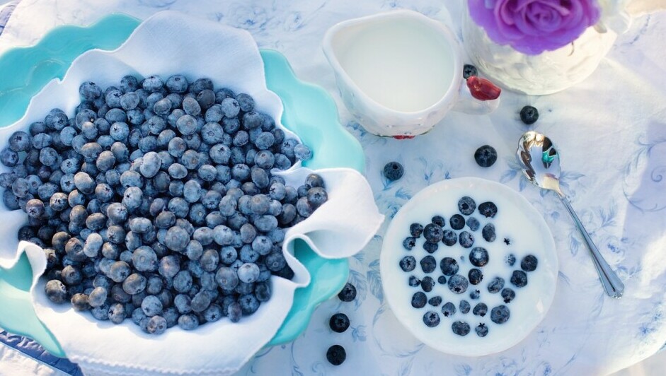 A bowl of enticing looking blueberries on a table next to a pot of fresh cream