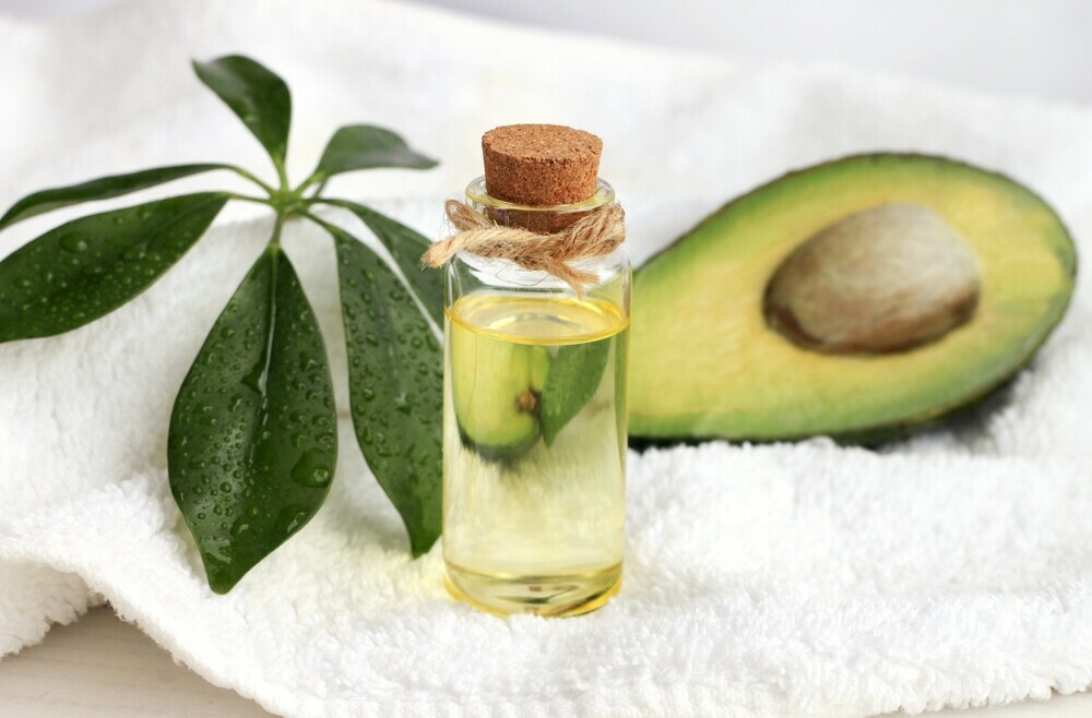 half avocado, bottle of avocado oil, and avocado leaves lying on white towel