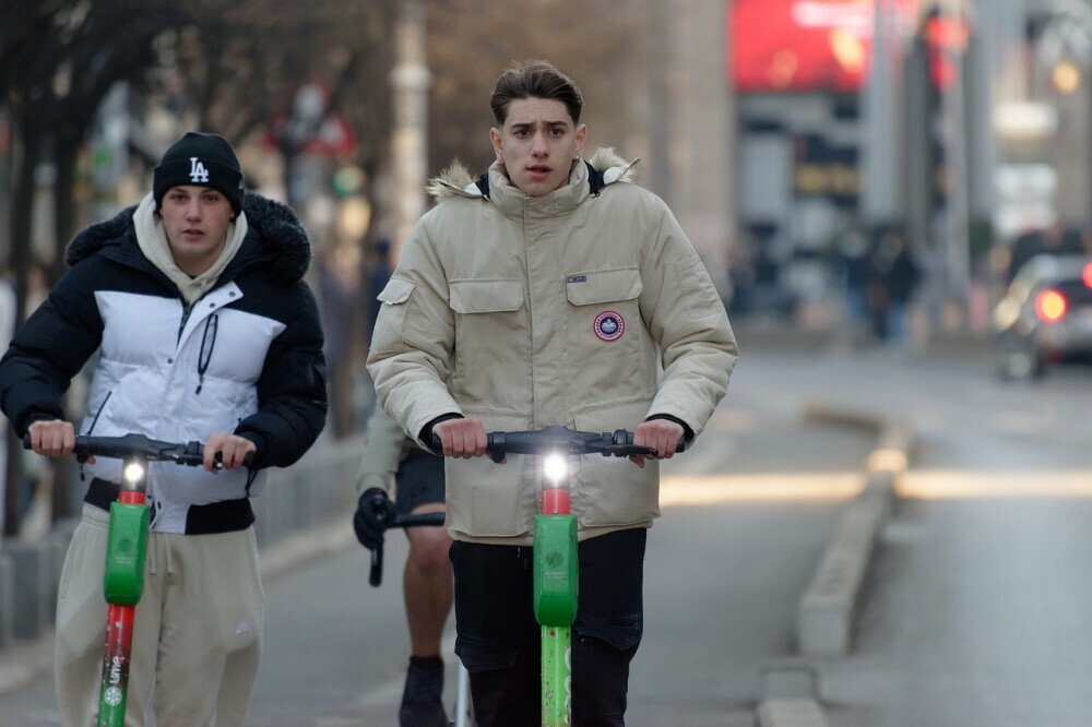 Two young individuals riding through the city on hired electric scooters without helmets with a cyclist behind 