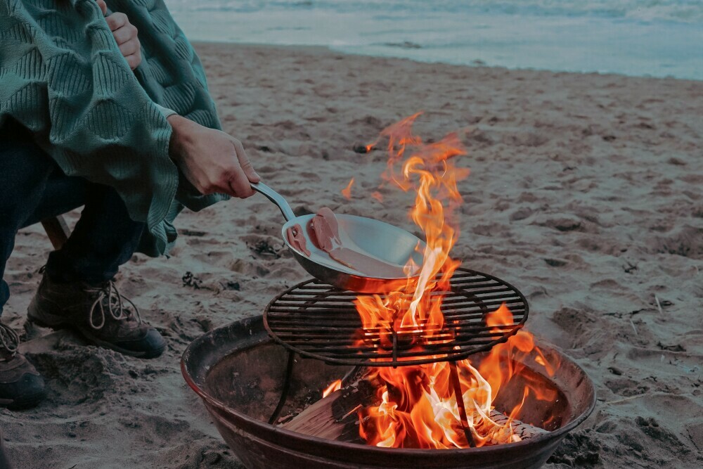 Bacon being cooked in a titanium frying pan over a fire pit on the beach