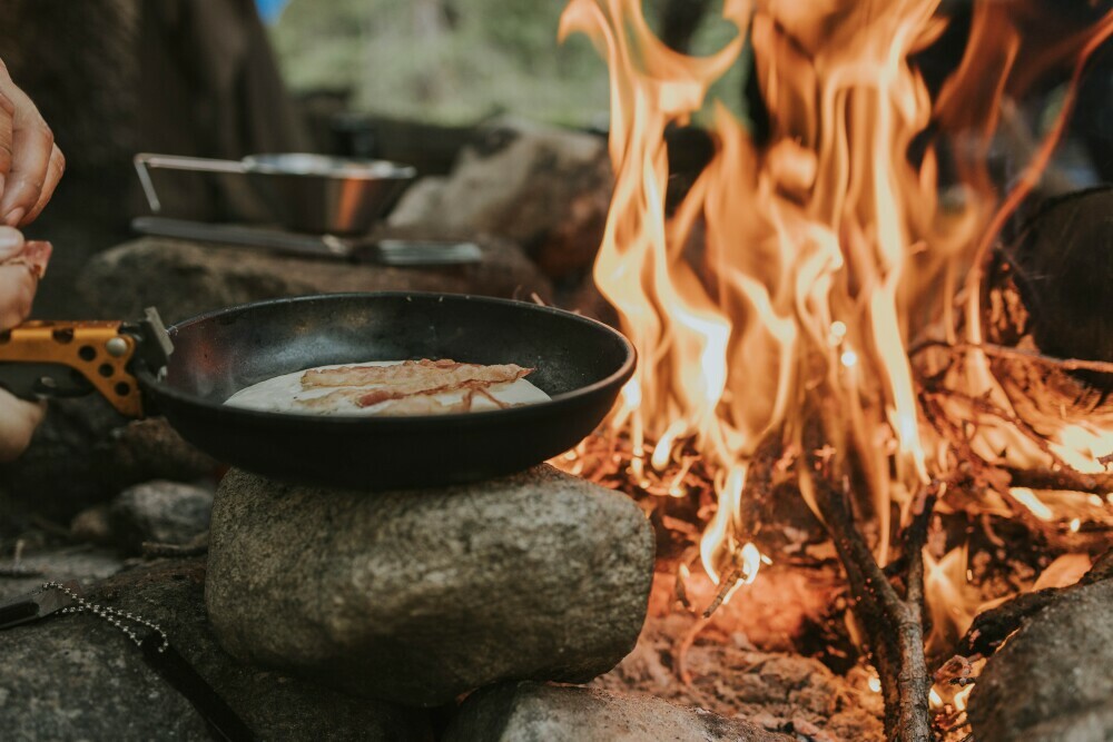 Sizzling bacon being cooked in a frying pan on a rock close to a roaring campfire