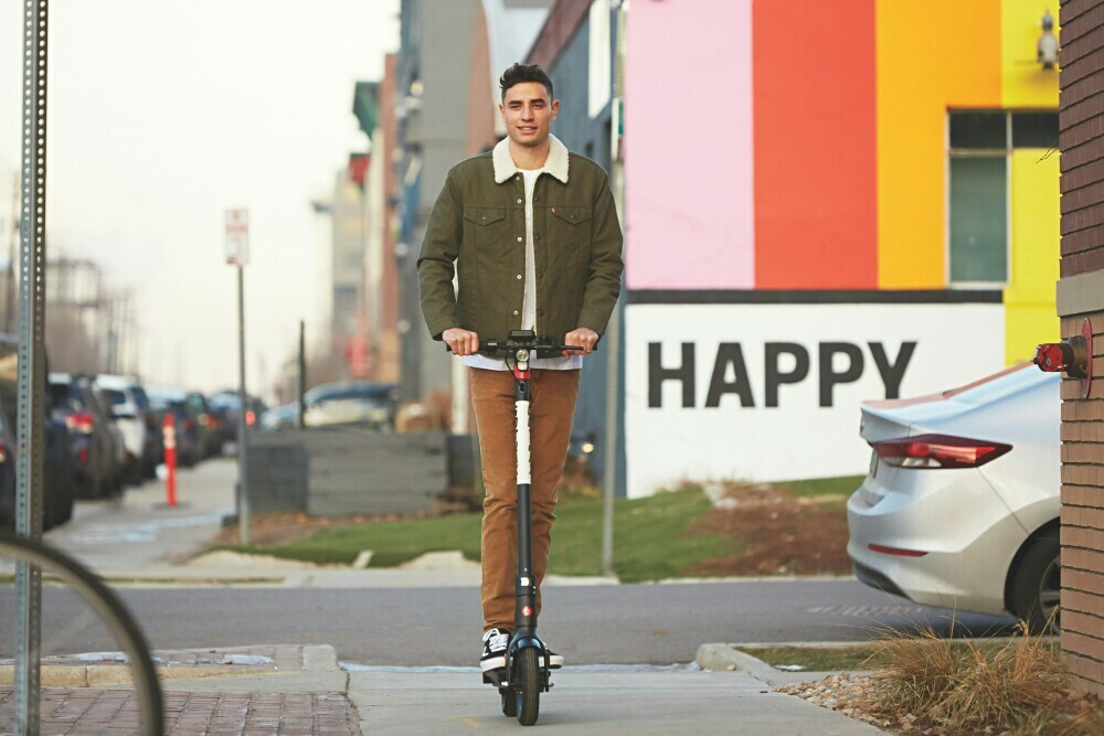 Individual riding his e-scooter along a pavement with cars and buildings all around and a sign reading "HAPPY"