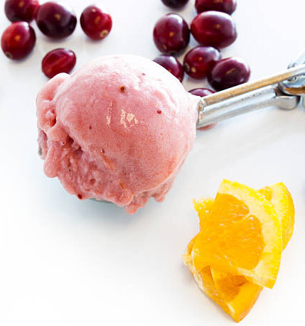 Homemade Cranberry Orange Ice Cream Recipe image 6 scoop of cranberry and orange ice cream in ice cream scoop with fresh cranberries and slices of fresh orange to the side white background frosted fusions