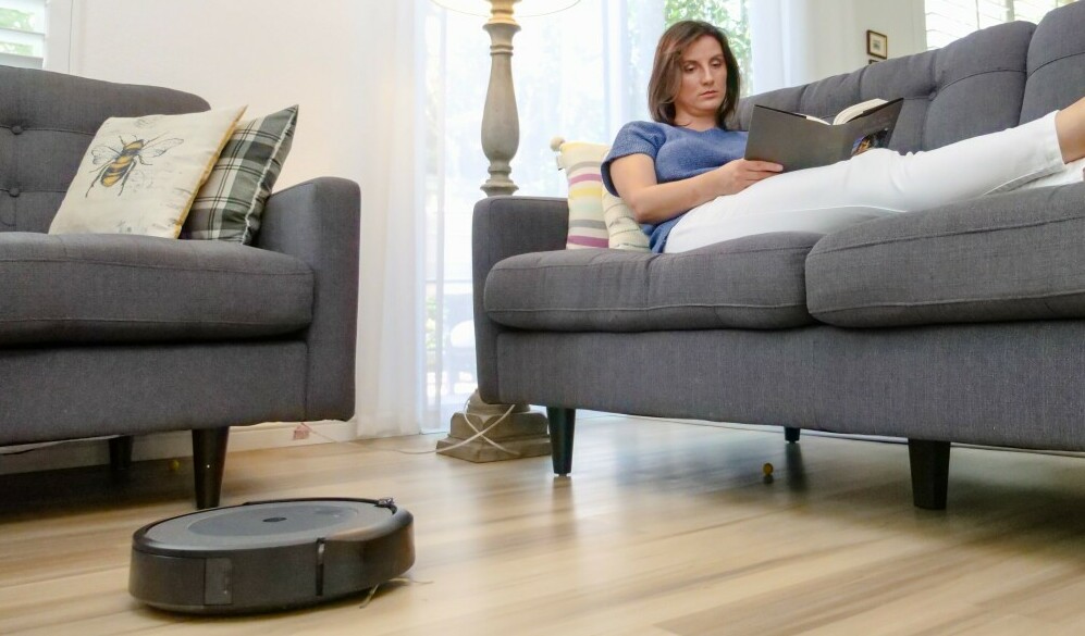 Roomba Robot Vacuum cleaning floor while relaxed woman on couch is reading a book.