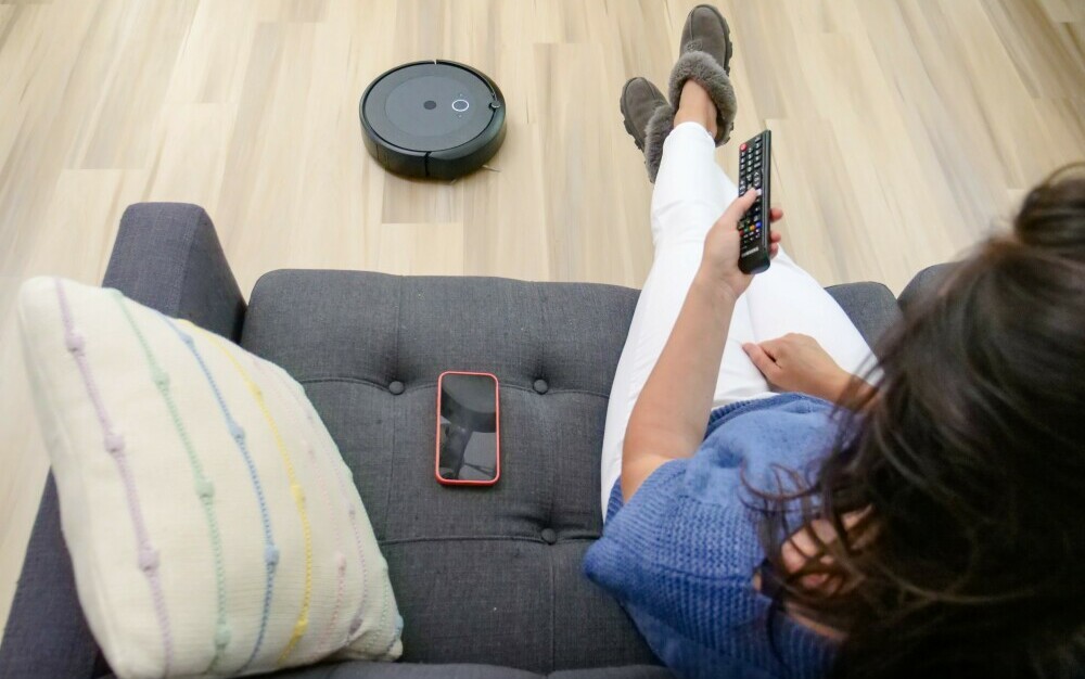 Roomba Robot Vacuum cleaning hardwood floor while woman relaxing on couch watches television.