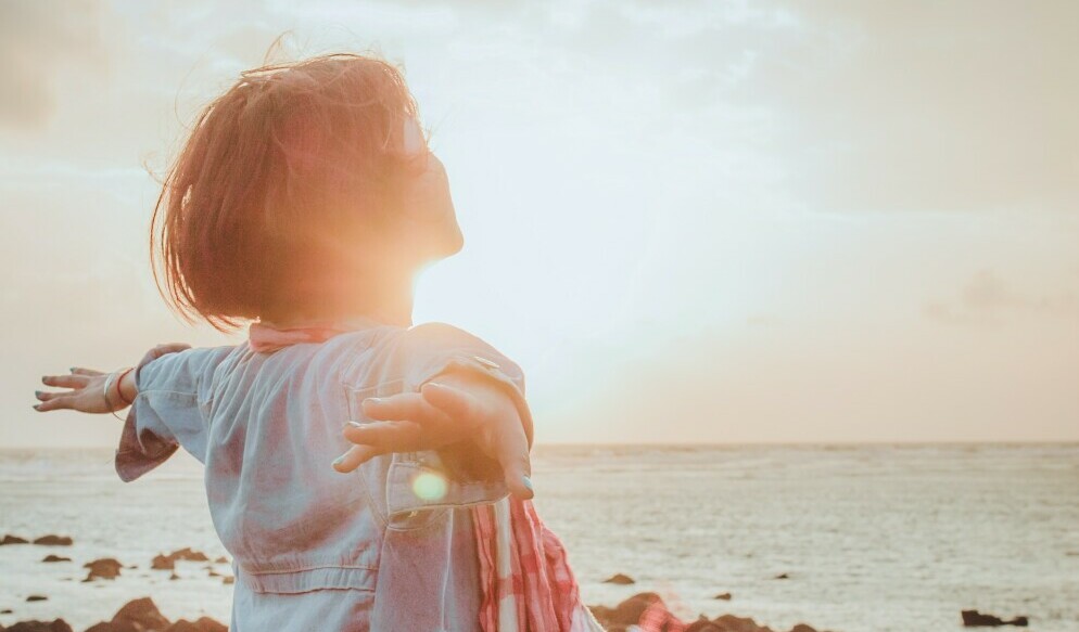 Woman standing on a beach with open arms and facing the direction of the sun