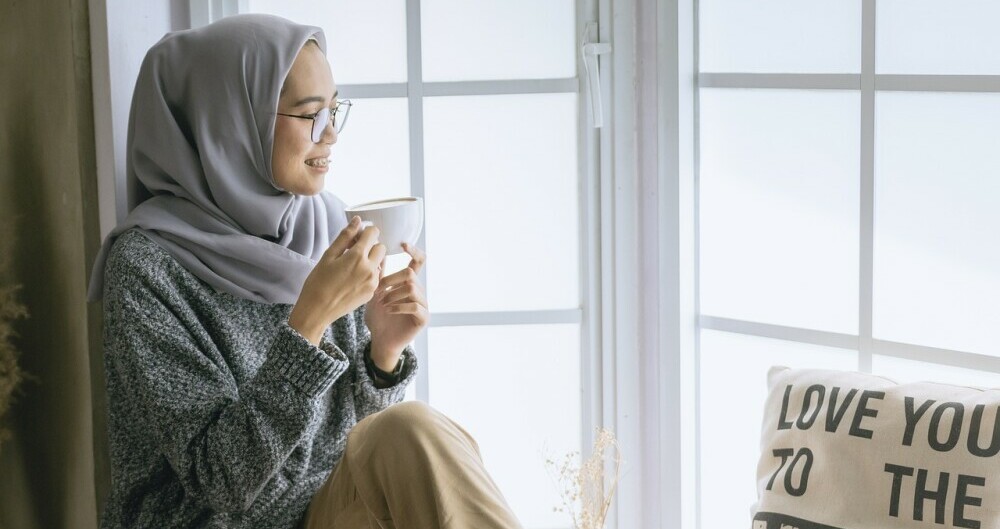 Young woman with a scarf drinking a cup of tea and looking out her window smiling