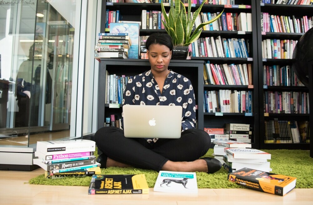 Girl sitting on a library floor with a stack books around and a laptop on her thighs