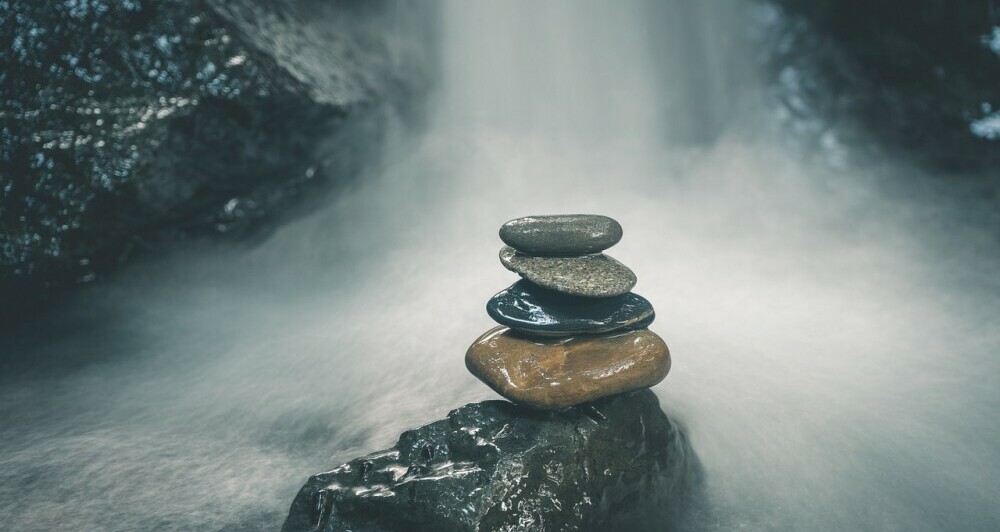 A large rock sitting in the middle of a pool where water from a waterfall is cascading down soaking the large rock. On top of the rock are four stones,, decreasing in size and stacked in a balancing pile. These rocks and stones are different colours