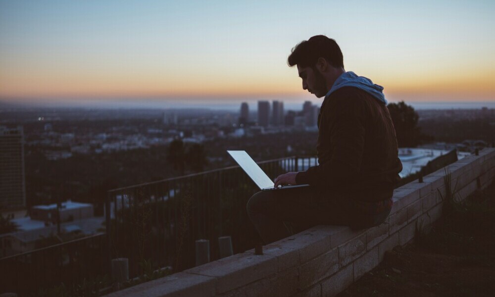 A wintry sun is setting over a distant city. In the foreground and almost in silhouette from the darkening sky is a lone individual, sitting on a low parapet wall working on a laptop computer  