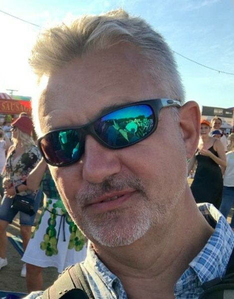 Picture of a Male wearing a blue and white check casual shirt which is open at the neck. The Male is also wearing sunglasses and the background shows other people indicating the picture was taken at an open air summer event such as a concert.