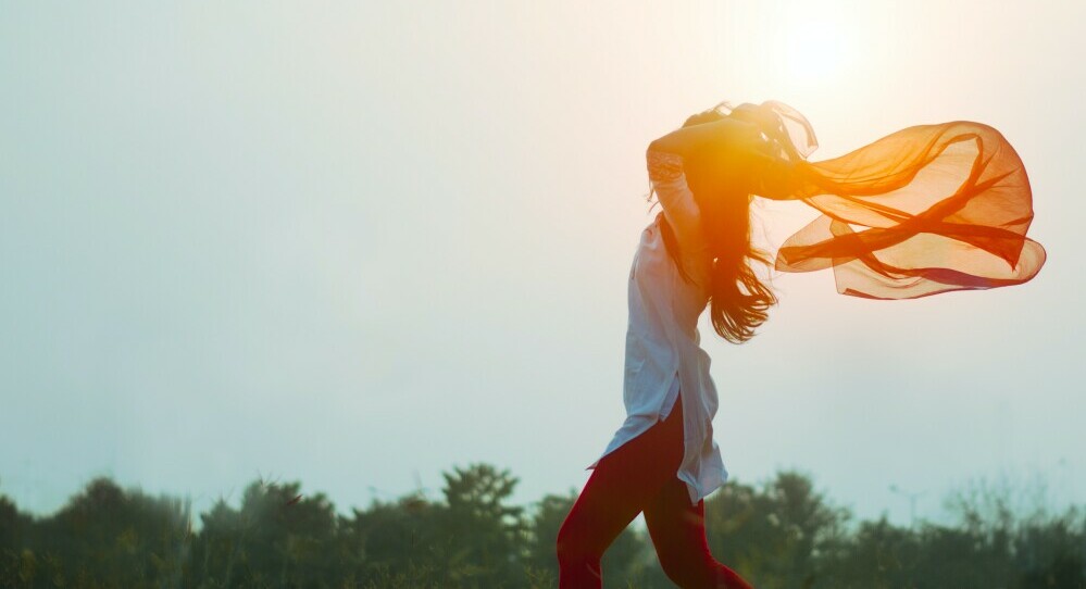 A hazy sunshine backdrop with blurred trees in the background. Standing to the right in the image is a girl with long hair, that is blowing in the breeze and to convey freedom the girl is holding above her head a long sheer orange scarf that is blowing in the breeze.