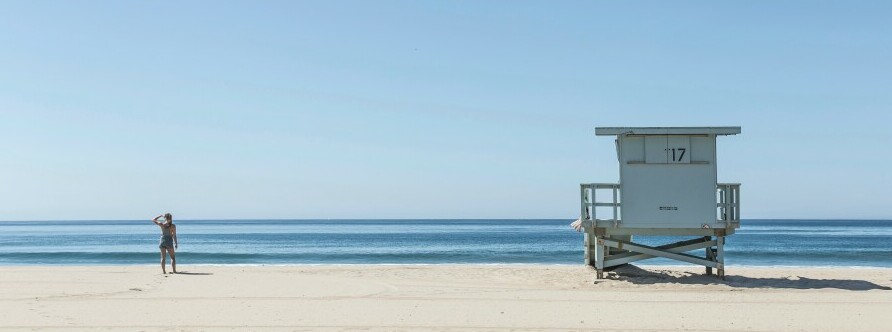 A Clear blue sky over a flat sand beach with a lone individual on the left and in the distance, shielding their eyes from the sun  whilst looking out to the sea