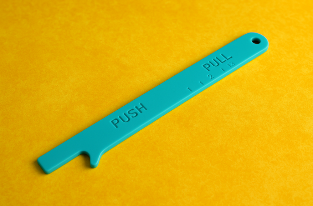 A turquoise silicone oven ruler lies diagonally across a bright yellow countertop, clearly showing its hook notch and embossed 