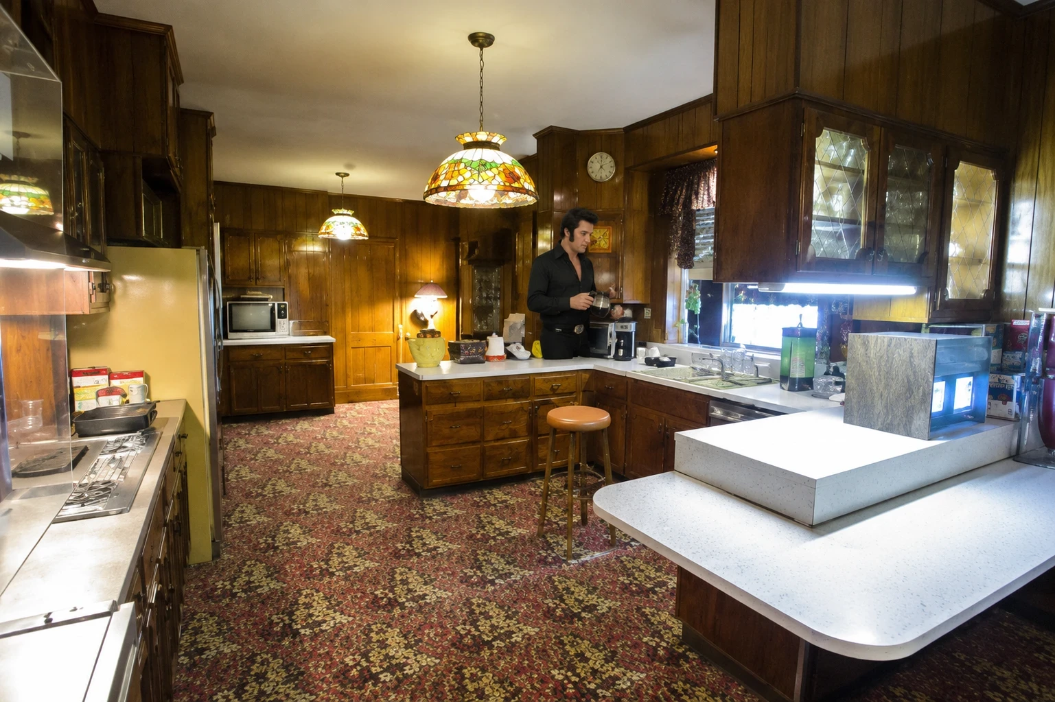 Elvis Presley standing in the Graceland kitchen surrounded by dark wood paneling, vintage lighting, patterned carpeting, and retro kitchen décor.
