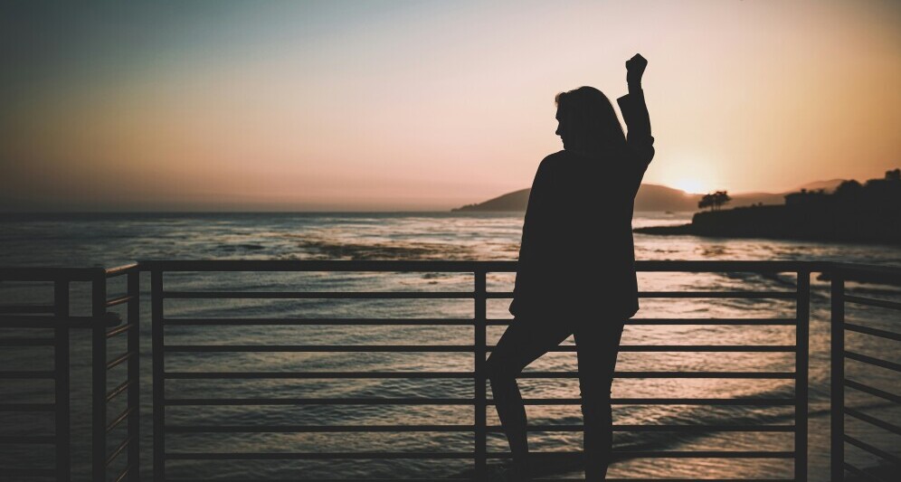 a woman standing on a deck behind a railing overlooking the ocean. she is fist pumping the air in triumph.