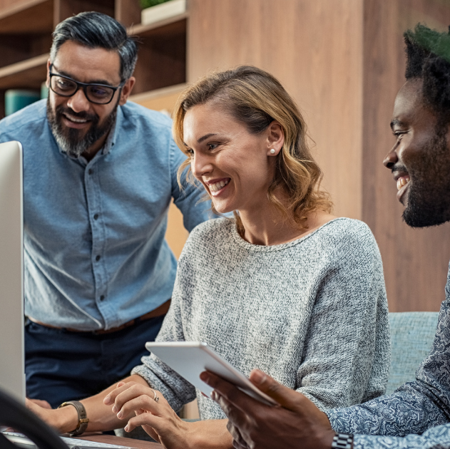 Three coworkers, a blonde woman in front of the computer, a bearded man with glasses on the left side and a man holding a tablet on the right side. All smiling.