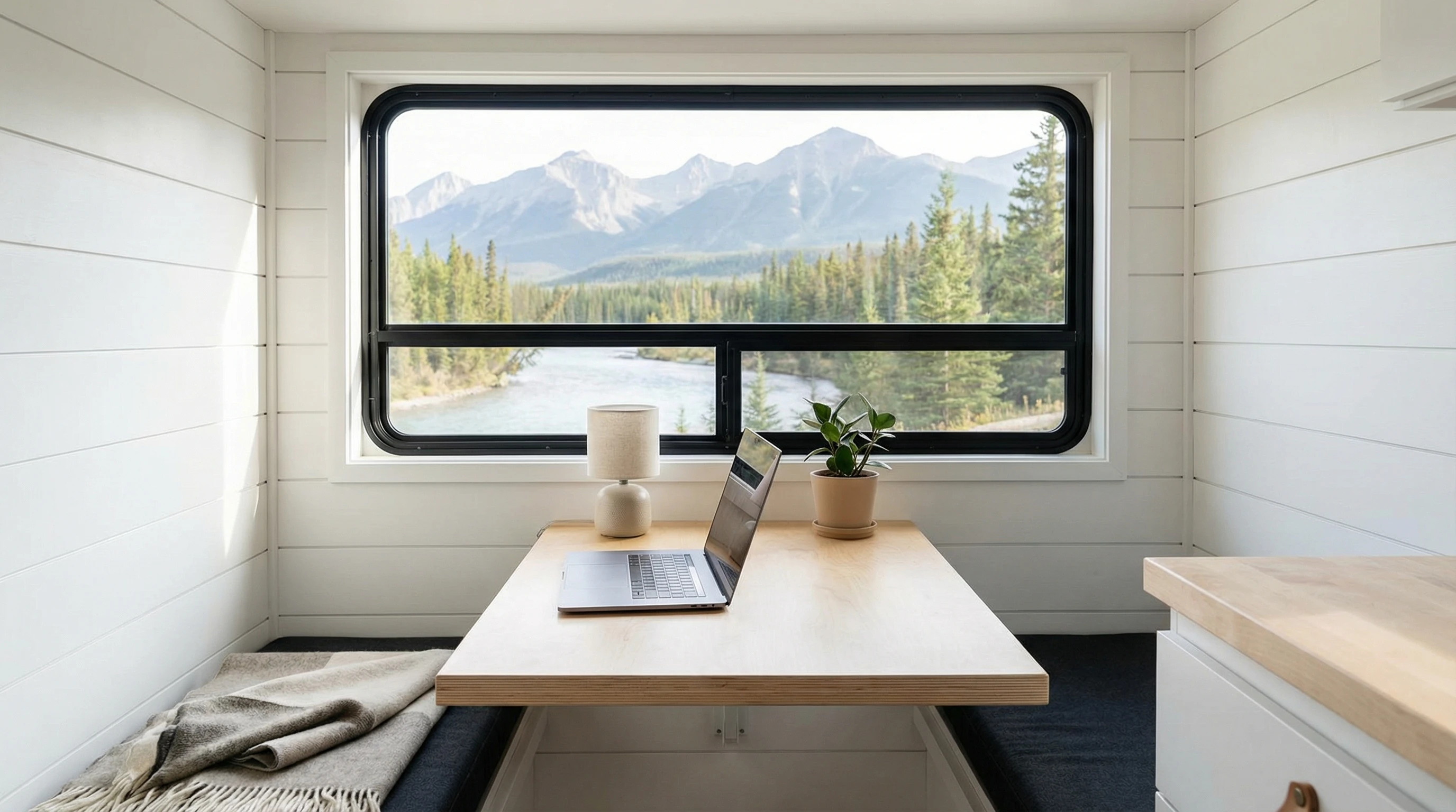 Minimalistic RV interior showing a workspace setup, including a small desk, laptop, lamp, and a backdrop of nature